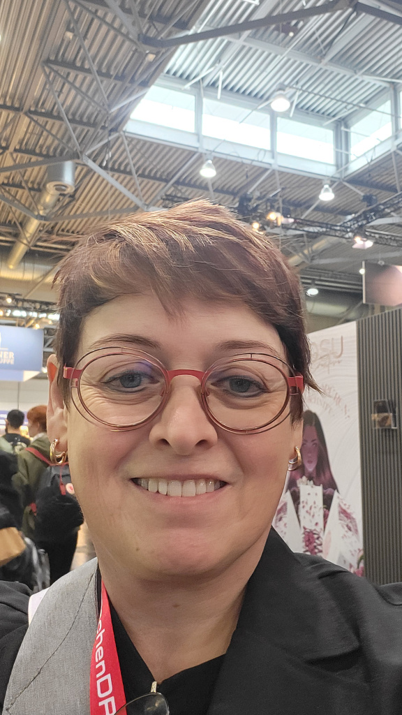 Smiling person with round red glasses in an exhibition hall with high ceilings and light trusses
